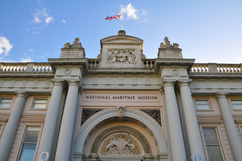 White facade of a building with several columns and the british flag flying from the top. The sign above the arched door reads National Maritime Museum. 