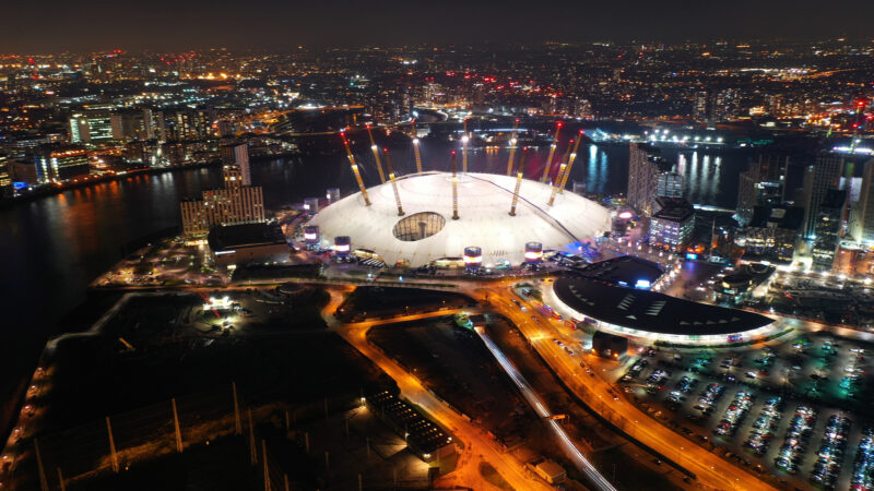 Aerial night shot above the O2 Arena, a large, white dome with numerous yellow poles rising from the top, surrounded by London city skyline.