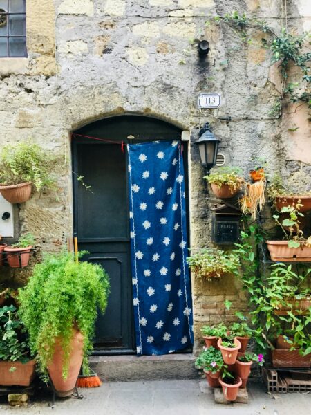 Doorway in a grey stone wall with a blue and white flower patterned rug hanging in front of the door and potted plants all around. 