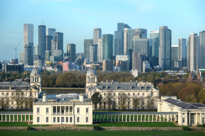 Large white palace or mansion with collonaded walkways either side and a view of London city skyline behind with many skyscrapers against a blue sky. 