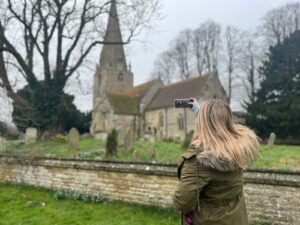 Emily wearing a khaki green parka with a fur lined hood facing away from the camera using a smartphone to take a photo of a church in England on a grey day