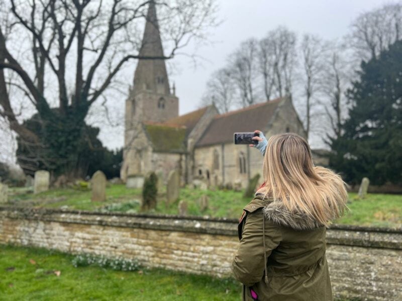 Emily wearing a khaki green parka with a fur lined hood facing away from the camera using a smartphone to take a photo of a church in England on a grey day. Orange Travel eSIM review. 