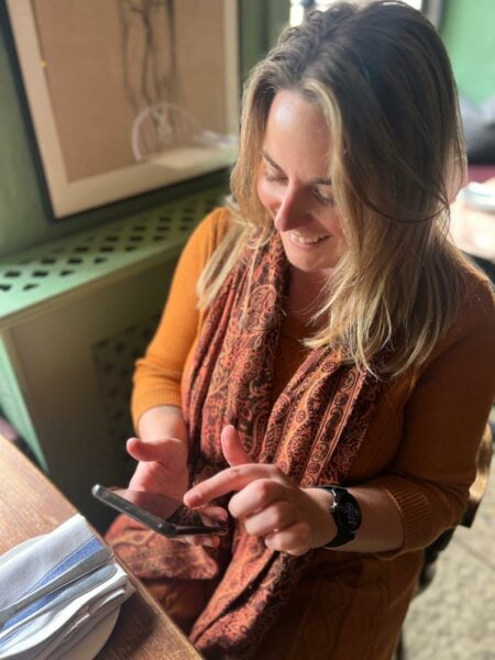 Emily wearing a mustard yellow dress and orange scarf sitting at a wooden table in a pub looking at her phone and laughing.