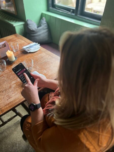 Looking over Emily's shoulder as she is sat at a wooden table in a pub changing the settings on her smartphone. She is wearing a mustard yellow dress. 